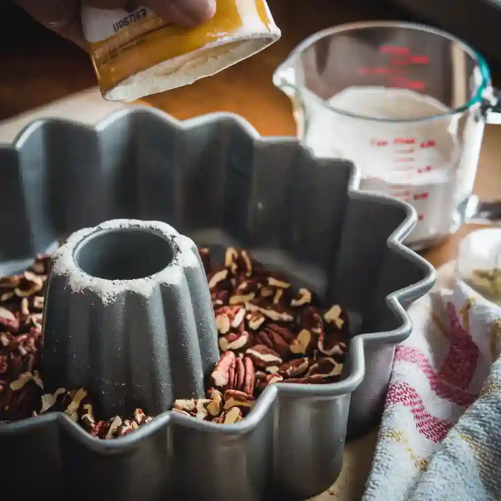 A well-greased Bundt pan being coated with butter and lightly dusted with flour. A small bowl of finely chopped pecans is being sprinkled at the bottom. A folded kitchen towel, a measuring cup of flour, and soft, golden lighting complete the cozy baking ambiance.