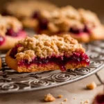 Close-up of freshly baked raspberry crumble cookies with a golden, buttery streusel topping and a vibrant raspberry filling, placed on a decorative cooling rack.