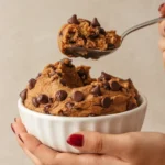 Delicious edible chocolate chip cookie dough in a white bowl, garnished with chocolate chips, held by a hand with a spoonful lifted.